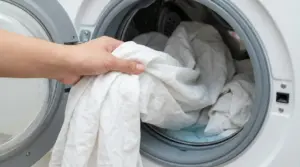 Una mano inserisce lenzuola bianche in una lavatrice aperta