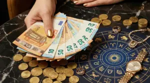 Mani con banconote in euro, monete e oggetti di lusso su una tavola con simboli zodiacali