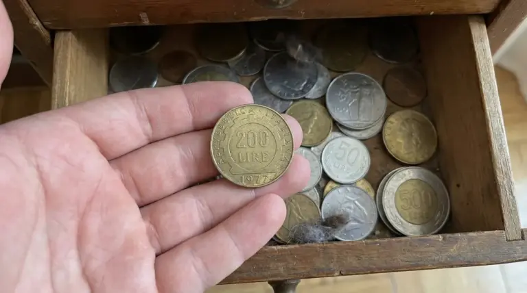Una mano tiene una moneta da 200 lire del 1977 sopra un cassetto pieno di vecchie monete italiane.